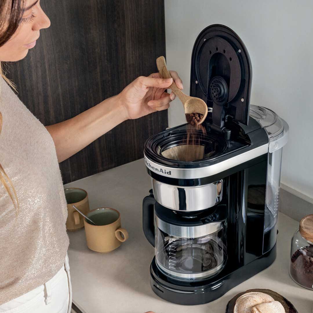Hölgy őrölt kávét tölt a fekete KitchenAid filteres kávéfőző aranyszűrőjébe